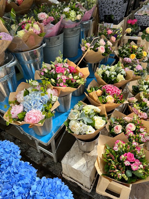A vibrant display of freshly arranged floral bouquets featuring pink, white, and pastel-colored roses, hydrangeas, and other seasonal flowers, all wrapped in brown craft paper and set in metal buckets