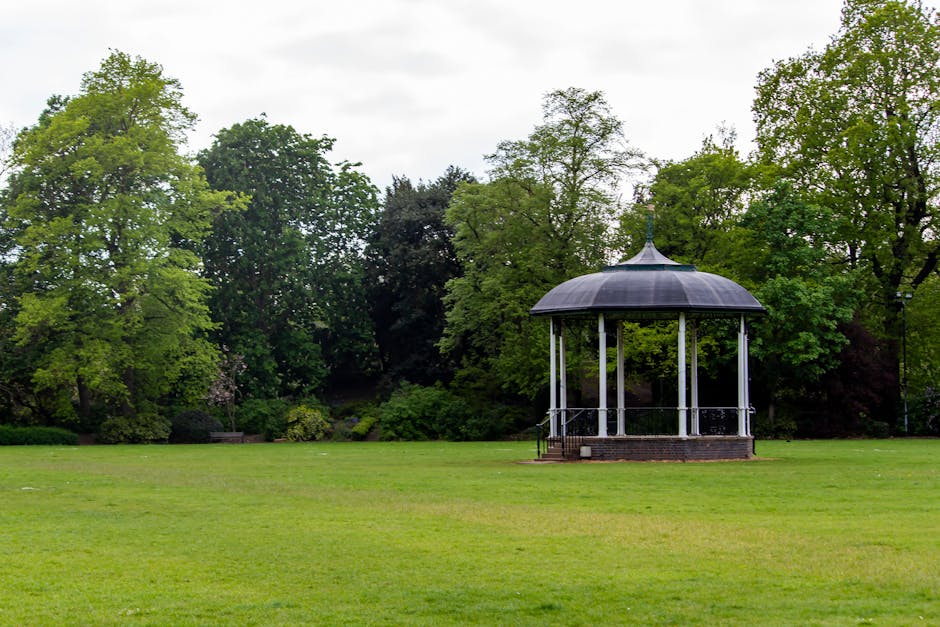 A black and white open-air bandstand with a domed roof situated on a grassy area in Wanstead Park. Surrounding the bandstand are lush, green deciduous trees with fresh spring leaves, creating a peacef